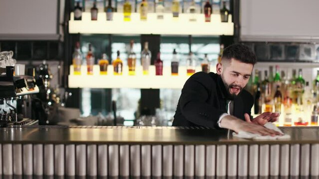 Caucasian bearded barman in uniform wiping wooden surface of bar counter with rag. Handsome man cleaning working place and preparing restaurant for clients.