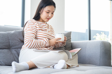 Young asian woman in casual clothes reading magazine, drinking tea on couch in modern interior