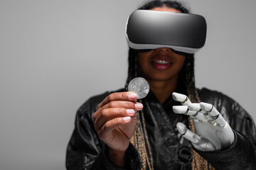Fototapeta na wymiar KYIV, UKRAINE - APRIL 28, 2022: Blurred african american woman with vr headset and robotic hand holding bitcoin isolated on grey.