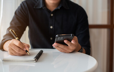 Male use a smartphone and take notes in the notebook at the desk in the morning.