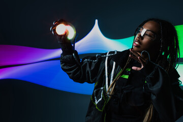 African american woman in smart glasses holding lights on grey background with abstraction.