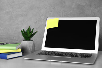 Fototapeta premium Laptop with empty sticky note and stationery on white table near grey wall. Space for text