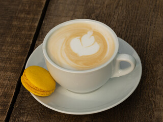 Cup of cappuccino with yellow macaron on wooden table. Close up
