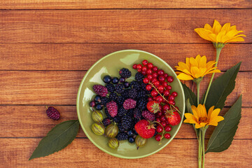 Berry mix - currants, gooseberries, mulberries, strawberries on a green saucer with yellow flowers on a wooden background with space for design. Summer concept.