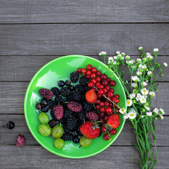 Berry mix - currants, gooseberries, mulberries, strawberries in a plate near a bouquet of wild flowers on a wooden background with copy space. Summer concept.