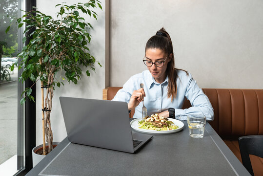 Young Freelancer Business Woman Watching Movie On Her Laptop Computer During Her Lunch Break. Exchange Student Waiting For Video Call With Her Family While She Eating Big Portion Of Chicken Dish