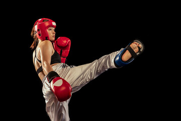 High kick. Attractive female boxer in boxing gloves and helmet training isolated on dark studio...