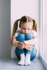 Little sad girl sitting on windowsill at home
