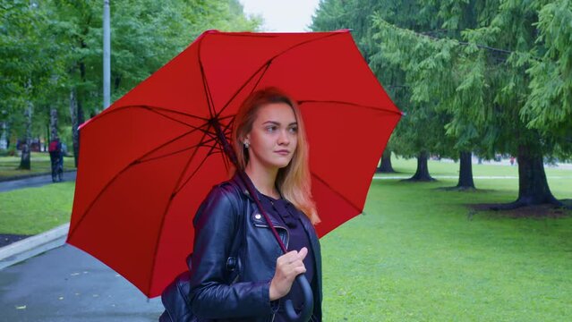 Beautiful girl with red umbrella stands at path in the park. Romantic blonde lady walking at alley in summer. Portrait of pretty young woman in leather jacket outside