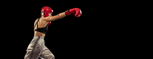 Young female boxer in boxing gloves and helmet training isolated on dark studio background. Sport, achievements, motivation, hobby, results, success concept - Powered by Adobe