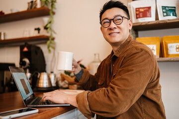 Adult asian man drinking coffee and using laptop in cafe indoors