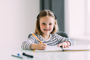 Close up photo of a little girl drawing something in her notebook sitting at the table in the kitchen