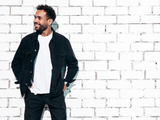 Handsome smiling hipster model. Sexy unshaven man dressed in black  summer shirt and jeans clothes. Fashion male posing near white brick wall in studio at sunny day. Isolated