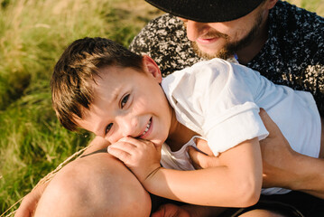 Father's Day. Happy son hugging dad and sitting on the green grass in the field in summer mountains. Dad and son playing at sunset. Kid with parents smiling, spending time together. Children's Day.