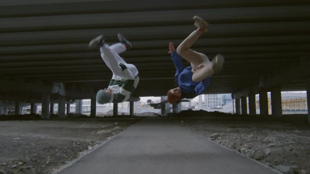 Following shot of two young athletes running on street under urban bridge and performing side flips in slow mo while practicing parkour stunts