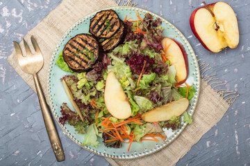 Healthy food apple salad with roasted eggplant red lettuce in plate has fork on grey wood table.
