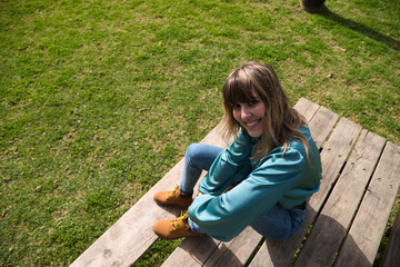 Naklejka na ściany i meble Young, pretty blonde woman dressed in jeans and boots is sitting at a wooden table in the park. In the background you can see the river on a sunny day. The woman is happy. Concept holidays and free.