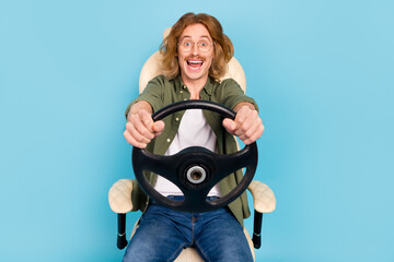 Portrait of attractive cheerful comic guy sitting in armchair holding steering wheel fooling isolated over pastel blue color background