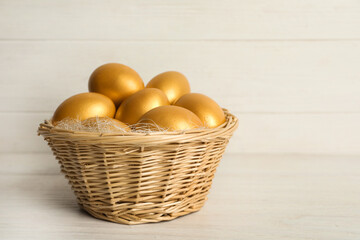 Shiny golden eggs in wicker basket on white wooden table, space for text