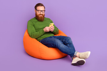 Full size portrait of cheerful satisfied man sitting bag hold use telephone isolated on purple color background