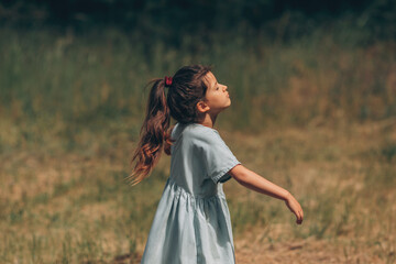 A little schoolgirl girl in a dress spread her arms to the sides in a nature meadow, she enjoys the open air and freedom