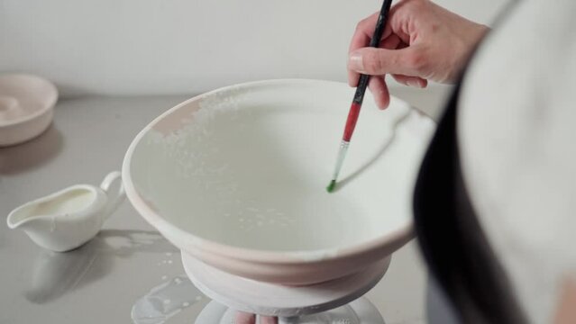 Female hands paint a ceramic bowl with a brush. Women's hands apply white paint on a blue base, the artist works in a pottery studio with a potter's wheel. Close up.