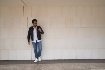 Handsome young man with beard, sunglasses, leather jacket, white shirt and jeans, leaning against a white wall, consulting his cell phone. Concept beauty, fashion, smartphone, app, social networks.