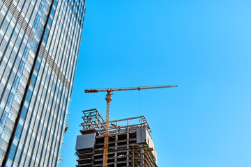 Fototapeta premium The base of a building under construction and a yellow tower crane along it next to part of a glass skyscraper against a clear blue sky in the light of the sun