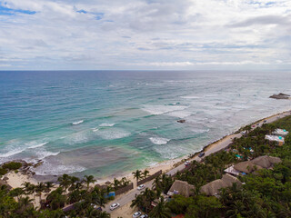 Aerial drone view of Tulum beach road surrounded by nature with wavy Caribbean Sea and white sand beach on a cloudy day 