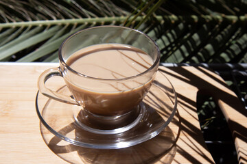 Glass cup with fragrant cappuccino coffee on a summer veranda against the backdrop of palm trees