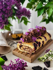 Fototapeta na wymiar Currant roll with chocolate cream, decorated with lilac flowers on a wooden stand with a cup of tea.