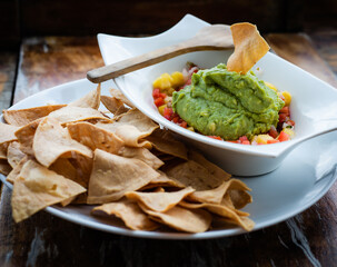 Mexican guacamole with mango, tomatoes and corn chips served in white plate with wood spoon