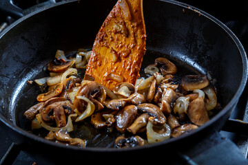 Mushrooms fried with onions in a deep frying pan with a wooden spatula.
