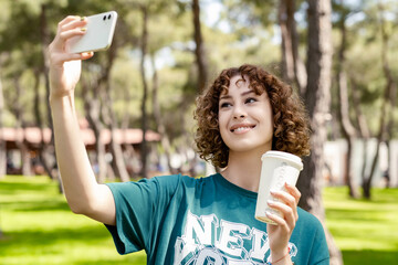 Cheerful redhead woman wearing green tee and standing on city park, outdoors taking selfie and holding takeaway coffee mug. Self portrait for social media. Outdoor sport concepts.