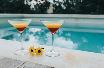 Orange and red cocktails with yellow flower next to swimming pool with water reflection in the jungle near Tulum at sunset