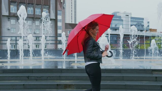 Happy girl in leather jacket walks down street past fountains, smiles coquettishly, twirls red umbrella in her hand. Attractive blonde woman enjoys every moment, flirts, is joyful of pleasant event