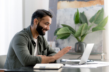 Online conversation. Positive successful Arabian or Indian man, sitting in his modern office, talking with colleagues or client remotely via video call by laptop, discussing a project, smiles friendly