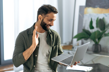 Successful arabian or indian man, holds and using laptop while stand in modern office, talking with coworker or client via video conference by laptop, conducts virtual meeting, smiling friendly