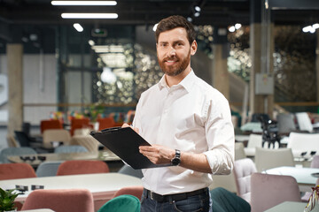Front view of brunette male with beard standing, holding folder in furniture store. Designer, seller, architect writing, designing, looking at camera. Concept of furniture. - obrazy, fototapety, plakaty