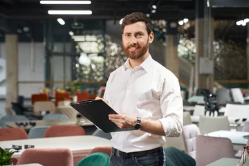 Foto op Canvas Front view of brunette male with beard standing, holding folder in furniture store. Designer, seller, architect writing, designing, looking at camera. Concept of furniture. © serhiibobyk