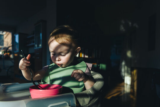 A baby wearing a bib eats out of a bowl in dramatic lighting