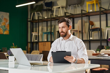 Front view of boy with beard sitting at table, designing. Handsome designer writing, using laptop,...
