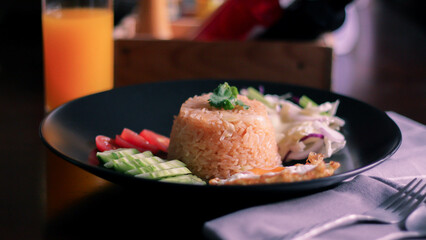 Fried rice, fried egg, cucumber, sliced tomato, green salad and Fried chicken placed on a black plate as a garnish.