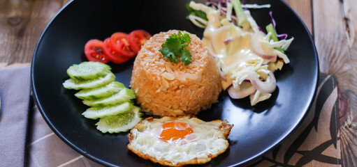 Fried rice, fried egg, cucumber, sliced tomato, green salad and Fried chicken placed on a black plate as a garnish.