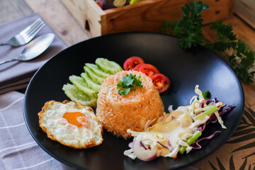 Fried rice, fried egg, cucumber, sliced tomato, green salad and Fried chicken placed on a black plate as a garnish.