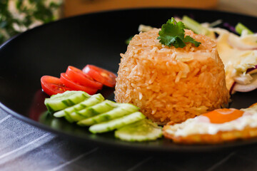 Fried rice, fried egg, cucumber, sliced tomato, green salad and Fried chicken placed on a black plate as a garnish.