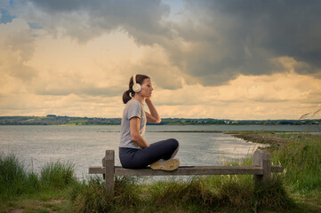 Fototapeta na wymiar Single woman sitting on a bench beside water listening to music on her headphones.