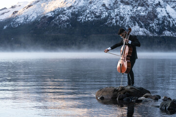 Musician playing cello by the lake with snow capped mountains on the back