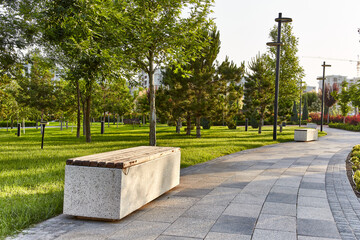 Obraz premium White stone and wood bench on the sidewalk in the park with fresh green grass and trees around at dawn light with houses in the background