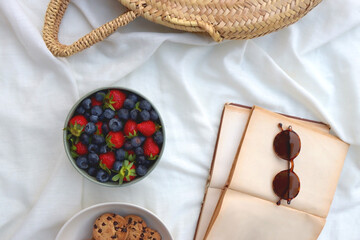 Picninc blanket with straw bag, bowl of strawberries and blueberries, bowl of chocolate chip...